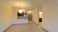 This single-floor residential space offers an open-concept design that seamlessly connects the kitchen and living area, creating a functional and cohesive layout. The kitchen is outfitted with dark espresso cabinetry, granite countertops, and stainless steel appliances, anchored by a breakfast bar that provides additional seating and serves as a natural divide between the kitchen and living space. Overhead, stylish pendant lighting illuminates the kitchen area, adding a modern touch to the warm ambiance. The living area features neutral carpet flooring and is bathed in soft, warm lighting, contributing to a comfortable and inviting atmosphere. A hallway leads toward what appears to be a bedroom and a bathroom, which is visible through an open doorway and mirrors the same dark cabinetry found in the kitchen, maintaining a consistent design language throughout. The entryway door is centrally accessible from the main living corridor, adding to the practical flow of the space. Neutral wall tones and clean lines throughout give the unit a polished, move-in-ready feel that appeals to a wide range of residents.