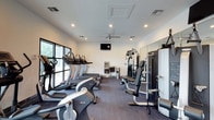 This fitness center offers a well-organized and fully equipped workout environment designed to support a wide range of exercise routines. The space is laid out on a single floor, making it easily accessible and straightforward to navigate.

Along one side of the room, a row of cardio machines — including treadmills and ellipticals — is positioned adjacent to large windows that allow natural light to fill the space and provide views of the surrounding greenery, creating an energizing atmosphere for workouts. The opposite side of the room features a multi-station strength training machine, offering a variety of resistance exercises, along with additional strength and functional training equipment.

The interior is finished with clean white walls and neutral-toned carpet flooring, contributing to a bright and focused environment. Recessed lighting is evenly distributed across the ceiling, supplemented by a ceiling fan that promotes airflow and comfort during intense sessions. A wall-mounted television is centrally positioned, providing entertainment or instructional content for users.

A water station is also visible within the space, adding a practical amenity for those exercising. The overall ambiance is modern, clean, and purposefully designed to accommodate both cardiovascular and strength training needs in one convenient location.