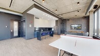 Located at 4555 N Sheridan Rd in Chicago's Uptown neighborhood, this contemporary single-floor amenity space combines modern design with functional versatility. The open-plan layout is anchored by an exposed concrete ceiling with industrial-style track lighting, creating a sophisticated urban aesthetic throughout.

The kitchen is a standout feature, outfitted with sleek blue lower cabinetry, white chevron-patterned upper cabinets, and a dramatic dark marble backsplash with bold veining. A large island with a dark countertop and plush blue velvet bar stools provides casual seating, while stainless steel appliances—including a full-size refrigerator and built-in oven—ensure full functionality. Light wood-look flooring runs throughout, adding warmth to the otherwise cool palette.

Adjacent to the kitchen, a stylish lounge area features a wall-mounted flat-screen TV set against decorative textured wallcovering, complemented by leather seating, a upholstered accent chair, and a wood media console. Floor-to-ceiling windows flood the lounge with natural light and offer expansive views of the surrounding Chicago cityscape. The space also includes glass-paneled entry doors and additional flexible areas suitable for gathering or events, making it a well-appointed communal destination.