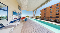 This two-floor waterfront property offers an impressive outdoor amenity experience defined by its architecturally distinctive rooftop pool deck. The centerpiece is a sleek lap pool set atop a broad composite wood deck, partially sheltered by a dramatic overhead canopy supported by angled concrete columns — providing shade without obstructing the sweeping views of the water and surrounding skyline. Adjacent to the pool, a generously sized lounge area features contemporary seating in bold accent colors, complemented by a built-in fireplace set against the building's light-paneled exterior facade. Large floor-to-ceiling windows and glass doors line the building's exterior, allowing abundant natural light and reinforcing a seamless indoor-outdoor connection. A separate open sun terrace provides rows of lounge chairs oriented toward the waterfront, ideal for maximizing unobstructed views of the bay. A long wraparound terrace on the lower level features smooth large-format tile paving, railing-framed waterfront vistas, and casual seating areas with direct access to the building's interior through multiple glass-panel entryways. Throughout the property, the design language is clean and contemporary, combining white paneling, dark-framed glazing, and refined outdoor materials that collectively create a resort-like atmosphere with a strong sense of place along the waterfront.