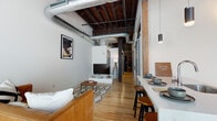 Located at 214 E Florida St in Milwaukee's vibrant Walker's Point neighborhood, this single-floor loft seamlessly blends industrial character with modern finishes. The open living area immediately captures attention with its soaring exposed wood beam ceiling, visible ductwork, and rich hardwood flooring — hallmarks of authentic loft-style architecture. Dark structural columns punctuate the space, adding to its raw, urban aesthetic.

The kitchen is well-appointed with dark cabinetry reaching to the ceiling, stainless steel appliances including a refrigerator, range, and microwave, complemented by a light quartz countertop and a subway tile backsplash. A bar-height counter with seating provides a casual dining area that flows naturally into the living space.

The bedroom offers a calm, neutral retreat with warm ambient lighting and a barn-style sliding door. Two bathrooms serve the unit — a full bath featuring stone-tile flooring and a walk-in shower with a white surround, and a half bath finished with a dark vanity, white countertop, and large mirror. Throughout, the contrast of crisp white walls against dark wood and matte black fixtures creates a cohesive, contemporary industrial ambiance ideal for urban living.