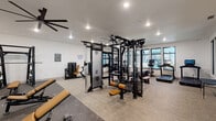 This modern fitness center is thoughtfully designed to support a full range of workout routines, offering a spacious, open layout with distinct zones for strength training, cardio, and free weights. The space features clean white walls, neutral-toned flooring with rubberized mat areas, and recessed lighting complemented by large, statement ceiling fans that promote air circulation throughout.

The strength training area is anchored by a robust Life Fitness multi-station cable and functional training system, alongside a power rack, adjustable benches with tan upholstery, and a dedicated dumbbell rack stocked with a full range of free weights. A heavy bag rounds out the functional training options, while a wall-mounted storage unit keeps accessories organized.

The cardio zone is lined along the windowed perimeter, featuring multiple treadmills with interactive digital displays, elliptical trainers, a stationary bike, a rowing machine, and a stair climber. Expansive black-framed windows run along two walls, allowing ample natural light to flood the space and offering views of the surrounding outdoor areas, including what appears to be a pool courtyard.

A separate, glass-partitioned room houses additional cardio equipment and provides a quieter workout environment. Restroom facilities are accessible directly from within the fitness center, adding convenience. The overall design is clean, contemporary, and highly functional.