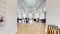 Located in Spring Hill, Tennessee, this thoughtfully designed community clubhouse offers a sophisticated, single-floor gathering space that blends comfort with modern elegance. The expansive open-plan lounge serves as the heart of the facility, featuring soaring tray ceilings, recessed lighting, and an array of decorative pendant and chandelier fixtures that create a warm, inviting atmosphere. Large multi-pane windows line the perimeter, flooding the interior with abundant natural light and framing views of the surrounding landscape. Light-toned hardwood flooring flows throughout, unifying the various zones seamlessly. A cozy lounge area anchors one side of the main room, complete with plush seating, a striking exposed brick accent wall, and curated artwork. Multiple high-top tables and communal dining arrangements encourage social interaction throughout the open space. The fully equipped kitchen features dark cabinetry, stainless steel appliances, quartz countertops, a stylish geometric tile backsplash, and a central island with bar seating — ideal for catering or casual gatherings. A private conference room, accessible through a glass-panel door, offers a refined setting with a rectangular chandelier, upholstered task chairs, wainscoting, and a glossy tile feature wall, making it well-suited for meetings or private events.