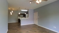 This single-floor space offers an open-concept design that seamlessly connects the living area and kitchen, creating a functional and inviting layout. The flooring throughout features wood-look tile laid in a classic pattern, providing both durability and a warm aesthetic.

The kitchen is well-appointed with dark cabinetry, stainless steel appliances, and a peninsula-style breakfast bar with seating, making it ideal for casual dining or entertaining. Pendant lighting above the bar adds a stylish focal point, while under-cabinet and recessed lighting ensure the cooking area remains bright and practical. A range hood is integrated above the stovetop, and a full-size refrigerator anchors the space.

The living area is open and airy, painted in a soft, neutral gray tone that complements the flooring and overall color palette. A ceiling fan with a light fixture is centrally installed, promoting airflow and providing additional illumination. A paneled entry door is visible along the wall, providing direct access to the unit.

The overall ambiance is clean, modern, and move-in ready, with a cohesive design that balances practicality with contemporary style throughout the single-level floor plan.