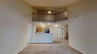 This two-story space is defined by its dramatic double-height ceilings that create an expansive, airy atmosphere in the main living area. The open-concept ground floor seamlessly connects the carpeted living space to a kitchen, which is accessible via a peninsula-style breakfast bar with a light-colored countertop. The kitchen is equipped with warm wood-toned cabinetry and black appliances, including a refrigerator. Recessed and flush-mount ceiling lights provide consistent illumination throughout the lower level. A doorway off the main living area leads to additional rooms, while a staircase on the right side of the space provides access to the upper level. The second floor features an open loft area enclosed by a classic wood balcony railing with vertical spindles, overlooking the living space below. A ceiling fan is mounted on the upper level, serving both floors with added air circulation. The neutral-toned carpet and beige walls throughout give the space a clean, cohesive look, while the vertical openness between floors adds a sense of grandeur to the overall design.