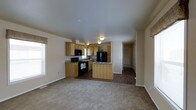 This single-story home offers a practical and well-organized open-concept floor plan that seamlessly connects the kitchen and living areas. The kitchen is a standout feature, showcasing light wood cabinetry that provides ample storage, granite-look countertops, and a full suite of black appliances including a refrigerator, gas range, and built-in microwave. A center island adds additional prep space and serves as a natural dividing point between the kitchen and the adjoining living area. The flooring transitions from dark hardwood-style in the kitchen and dining zone to plush carpeting in the living area, creating a warm and comfortable atmosphere throughout. Neutral beige tones on the walls contribute to a bright, clean aesthetic, while flush-mount ceiling lighting provides consistent illumination across the space. A window along the side wall allows natural light to filter into the living area. A hallway visible from the main living space leads to additional rooms, suggesting a well-connected and efficiently designed floor plan. The overall ambiance is cozy and functional, ideal for everyday living.
