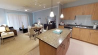 This single-floor residence showcases an open-concept layout that integrates the kitchen, dining, and living areas into one cohesive, flowing space. The kitchen is a standout feature, anchored by a substantial granite-topped island with warm honey-toned wood cabinetry and modern hardware. Pendant lighting hangs above the island, providing focused task illumination while adding a contemporary design element. The kitchen is further equipped with matching wood cabinetry along the perimeter, a dark countertop workspace, and stainless appliances including a range. Crown molding runs along the ceiling throughout, adding a refined architectural touch. The dining area sits adjacent to the kitchen and accommodates a round wooden table with modern white chairs, lit by track lighting overhead. Beyond the dining space, the living room opens naturally, featuring a ceiling fan, neutral-toned carpeting, and vertical blinds that allow for adjustable natural light. The walls are painted in a soft blue-gray tone that complements the warm wood and granite elements throughout. The overall ambiance is clean, modern, and well-appointed.
