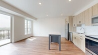 This thoughtfully designed one-bedroom, one-bathroom residence offers a bright and contemporary living experience across a single floor. The open-concept kitchen and living area serves as the heart of the home, featuring warm light wood cabinetry, stainless steel appliances, a white subway tile backsplash, light quartz countertops, and a central island. Light hardwood flooring runs throughout the main living areas, reflecting natural light and enhancing the airy, modern atmosphere. A sliding glass door in the living room opens to a private balcony with scenic outdoor views, while additional windows allow sunlight to pour in throughout the day. A wood-accented column detail adds a subtle architectural touch at the kitchen's edge. The bedroom offers plush carpeting, a ceiling fan, and a well-positioned window with views of the surrounding cityscape, creating a comfortable and restful retreat. A dedicated closet provides convenient storage. The bathroom is finished with a clean, minimalist aesthetic, including a wood-tone vanity, white countertop, framed mirror, and hardwood-style flooring. Recessed lighting throughout the unit completes the polished, contemporary feel of this well-appointed residence.