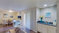 This single-floor residence offers a thoughtfully designed layout that balances functionality and comfort throughout. The open living area serves as the heart of the home, featuring durable wood-look flooring, a textured ceiling, and a sliding glass door with vertical blinds that fills the space with natural light and provides access to an outdoor area. A built-in wet bar with dark granite-style countertops and white cabinetry adds a distinctive touch, while a built-in shelving unit nearby offers practical display and storage space.

The galley-style kitchen is efficiently arranged with white cabinetry, dark countertops, black appliances including a refrigerator and electric range, and a stainless steel sink — providing ample workspace for cooking. Adjacent to the kitchen, a cozy dining nook with a textured ceiling offers an intimate setting for meals.

The bedrooms feature the same wood-look flooring, neutral gray walls, and ceiling fans for year-round comfort. One bedroom includes a window dressed with vibrant teal curtains that bring in natural light. The bathroom is finished with white cabinetry, dark countertops, and a tub-shower combination, maintaining the cohesive aesthetic found throughout the residence. Overall, the space presents a clean, contemporary feel with a consistent neutral palette accented by warm décor touches.