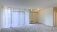 This single-floor residential space offers a straightforward and functional layout designed for comfortable everyday living. The main living area is a standout feature, characterized by large floor-to-ceiling windows dressed with vertical blinds that allow an abundance of natural light to fill the room. The soft, neutral-toned wall-to-wall carpeting adds warmth underfoot and contributes to the overall calm, understated aesthetic of the space. The walls are finished in a clean off-white tone, creating a bright and airy atmosphere that feels open and welcoming. A flush-mount ceiling light provides overhead illumination, complementing the natural light during evening hours. The room also includes a built-out overhead soffit detail that adds a subtle architectural element to the space. Ventilation is well-considered, with both ceiling and floor-level HVAC registers visible, ensuring efficient climate control throughout. The overall ambiance is clean, neutral, and move-in ready, providing an excellent blank canvas for personalization. The single-floor configuration ensures easy accessibility and a cohesive flow throughout the living space.