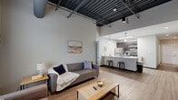 Located at 2611 E Third St in Bloomington, Indiana, this modern 4-bedroom, 3-bathroom unit delivers a compelling blend of industrial design and contemporary comfort. The open-concept living and kitchen area is the heart of the unit, anchored by dramatically high ceilings with exposed steel beams and ductwork that lend a distinctive loft-style character. Large windows allow natural light to fill the space, complementing the warm wood-style plank flooring found throughout the common areas. The kitchen features stainless steel appliances, including a refrigerator, range, and microwave, paired with dark cabinetry and a sleek white countertop island with bar seating — ideal for dining or casual gatherings. A hallway with recessed lighting connects the shared spaces to the four bedrooms, each carpeted and designed to accommodate a bed and workspace. Select bedrooms benefit from the soaring exposed-ceiling aesthetic, while others offer a more intimate scale. The three bathrooms are clean and functional, each featuring a tub-shower combination and light wood-style tile flooring. A dedicated laundry area and four closets add practical convenience. This well-appointed unit offers a cohesive, modern aesthetic suited for shared living in Bloomington.