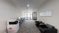 Located at 1005 S School Ave in Fayetteville, Arkansas, this contemporary single-floor office space is designed to support a range of professional and collaborative activities. The layout is thoughtfully organized, blending open-concept functionality with defined work zones.

The main area features a dedicated computer lab with a row of desktop workstations and ergonomic mesh chairs, accompanied by a commercial-grade printer and copier station. A sleek glass-and-black-steel partition wall separates the lab from an enclosed conference room, which benefits from generous natural light through exterior windows, creating a bright and focused meeting environment.

Adjacent to the workstation area is a private office nook outfitted with a round desk and task chair, ideal for individual work. The lounge zone provides comfortable black leather seating and accent tables, offering a welcoming space for informal meetings or waiting.

Throughout the space, patterned carpet tiles in neutral tones with teal accents add a modern, polished aesthetic that ties the zones together cohesively. Recessed lighting illuminates the interiors evenly, while contemporary artwork on the walls contributes to a professional yet approachable ambiance. The overall design balances productivity and comfort, making it well-suited for collaborative workgroups, academic support services, or professional office use.