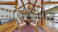 Located at 11 Parklands Dr in Bluffton, SC, this single-floor clubhouse delivers an impressive blend of rustic craftsmanship and modern amenity. The centerpiece of the space is a dramatic vaulted ceiling supported by rich, exposed timber beams and trusses, illuminated by cascading chandelier fixtures that lend a warm, lodge-inspired ambiance throughout. Wide-plank hardwood floors run continuously across the entire interior, grounding the open-concept layout.

The main gathering area features a floor-to-ceiling stone fireplace with a reclaimed wood mantel, flanked by shiplap accent walls that add texture and character. Comfortable seating arrangements, including leather club chairs and Persian-style area rugs, invite relaxed conversation and leisure. A billiards table anchors the game area nearby, complemented by additional entertainment features and vintage signage accents.

Large glass garage-style doors open fully along one wall of the dining area, seamlessly connecting the interior to an outdoor deck and allowing natural light and fresh air to flood the space. Multiple dining tables are positioned to take advantage of these outdoor views. Additional seating alcoves with high-top tables and mounted displays offer flexible gathering options. The overall atmosphere is welcoming, refined, and purposefully designed for community use and social engagement.