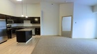 This single-floor space showcases an open-concept layout that connects the kitchen and living area in a clean, functional design. The kitchen is outfitted with dark espresso cabinetry, granite countertops, a stainless steel dishwasher, and an undermount sink. A kitchen island with matching dark cabinetry provides additional prep and storage space, while pendant lighting hangs overhead, adding a modern touch. The flooring in the kitchen and entry area consists of light hardwood, which transitions into plush neutral carpeting in the living space, creating a warm and comfortable atmosphere. Tall ceilings enhance the sense of openness throughout, and the neutral cream-toned walls provide a versatile backdrop. A doorway visible from the main living area leads to an additional carpeted room, suggesting a bedroom or secondary space beyond. The overall design is contemporary and understated, combining practical finishes with an airy, open feel suited for everyday living.