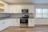 Located at 107 Wartburg Ave in Copiague, NY, this single-floor unit has been thoughtfully renovated with contemporary finishes and a clean, cohesive design. Light-toned hardwood flooring flows consistently throughout the space, complementing the neutral gray-white walls and bright white trim.

The kitchen is a standout feature, outfitted with crisp white shaker-style cabinetry, granite countertops, and a classic subway tile backsplash. A full suite of stainless steel GE appliances includes a refrigerator, gas range, over-the-range microwave, and dishwasher, providing a fully functional and modern cooking space. The kitchen opens toward a windowed dining or living area, allowing natural light to filter in through double-pane windows fitted with blinds.

The living areas are generously proportioned, offering an open and airy feel enhanced by the light flooring and neutral color palette. Baseboard heating units are present throughout, ensuring comfortable climate control. Additional rooms provide flexible living spaces, each featuring the same hardwood-style flooring and well-maintained finishes. The overall layout is practical and well-connected, with clear sightlines between rooms. The unit presents a clean, move-in-ready condition with a modern aesthetic suited for comfortable everyday living.