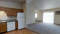 This single-floor residence offers a practical and comfortable open-concept layout that seamlessly connects the kitchen and living area. The kitchen is equipped with warm-toned wooden cabinetry, dark countertops, a full-size white refrigerator, and a dishwasher, all set atop easy-to-maintain hardwood-style flooring. The kitchen opens directly into a spacious living area, which features plush wall-to-wall carpeting and is bathed in natural light streaming through large windows fitted with vertical blinds. A baseboard heater along the living room wall provides efficient climate control throughout the cooler months. The transition between the hardwood-style kitchen floor and the soft carpeting in the living space creates a clear and functional delineation between the two areas. The neutral, light-toned walls throughout lend the interior a clean, airy feel, while the open sightlines between rooms enhance the overall sense of space. The vaulted ceiling visible in the kitchen area adds architectural interest and contributes to an open, expansive atmosphere. Overall, this residence offers a well-organized, functional layout suited for comfortable everyday living.
