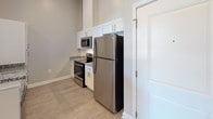 This single-floor residence offers a thoughtfully designed open-concept layout that seamlessly connects the kitchen and living area. The kitchen is outfitted with white shaker-style cabinetry, granite countertops, and a full suite of stainless steel appliances, including a refrigerator, range, and built-in microwave. A peninsula counter with a granite top provides a natural transition into the living and dining space, which features wood-look plank flooring, neutral gray walls, and a ceiling fan for added comfort. Natural light filters in through tall, vertically oriented windows that frame views of the surrounding exterior, contributing to an airy, open ambiance throughout. The bedrooms continue the same warm, neutral palette with matching plank flooring and generous wall space, each benefiting from the same tall windows that enhance the sense of height and openness. The bathroom is finished in a clean, all-white aesthetic with large-format tile flooring, a tub-shower combination, and simple, functional fixtures. Throughout the residence, clean white trim, panel-style doors with brushed nickel hardware, and consistent finishes create a cohesive, contemporary feel that is both functional and visually appealing.