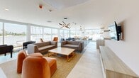 Located on S Sepulveda Blvd in Los Angeles, this contemporary rooftop lounge offers a well-appointed communal living space designed with both comfort and style in mind. The interior living area features warm terracotta and blush-toned walls, complemented by light tile flooring and a striking starburst chandelier that anchors the space. Comfortable seating arrangements, including plush gray and rust-orange sofas paired with a natural wood coffee table, create an inviting atmosphere for gathering and relaxation. A wall-mounted flat-screen TV with floating shelves and media storage adds a functional focal point to the lounge.

Floor-to-ceiling sliding glass doors flood the interior with abundant natural light and frame sweeping panoramic views of the Los Angeles skyline and surrounding palm-lined cityscape. These doors seamlessly connect the interior to a generous wraparound outdoor terrace finished in large-format stone tile. The terrace offers built-in planters with lush greenery, additional seating, and unobstructed open-air views under the Southern California sun, making it an ideal space for both casual lounging and social events. The overall design balances modern minimalism with warm, welcoming touches throughout.