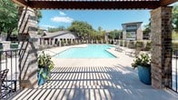 Located at 6401 Hulen Bend Blvd in Fort Worth, Texas, this outdoor amenity area is a well-appointed community retreat designed for relaxation and social gatherings. The centerpiece is a generously sized swimming pool with clear, turquoise water, surrounded by a spacious concrete deck furnished with lounge chairs and sun loungers, ideal for soaking up the Texas sunshine.

A handsome wooden pergola frames one side of the pool area, casting a pleasing pattern of light and shadow across the deck. Beneath it, multiple round patio tables with wrought-iron chairs and vibrant red umbrellas provide comfortable shaded seating for outdoor dining and socializing. The pergola's structured design adds an architectural element that complements the surrounding brick and stone buildings.

The space is enclosed by a decorative iron fence with stone pillar accents, lending both security and visual cohesion to the area. Lush, mature trees create a natural canopy that softens the environment and provides additional shade throughout the day. Views of the surrounding multi-story residential buildings and a well-maintained lawn with a visible volleyball net area extend beyond the pool enclosure, contributing to the community-oriented atmosphere. The overall ambiance is inviting, combining thoughtful design with practical outdoor amenities in a lush, green setting.