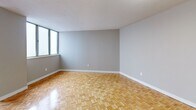 Located at 1 Dean Park Rd in Toronto's east end, this three-bedroom, single-floor unit offers a practical and well-organized layout. Throughout the space, warm parquet flooring in a classic basket-weave pattern adds character and a timeless aesthetic to every room.

The living area is generously proportioned, featuring neutral grey walls and a pass-through kitchen window that connects it to the kitchen beyond, where appliances including a refrigerator are visible. This open sightline creates a sense of connectivity between the living and cooking spaces.

The spacious primary bedroom is notably large, offering ample room for furnishings, and is bathed in natural light through a large sliding window. The two additional bedrooms are comfortable in scale, each with the same parquet flooring and neutral tones. A dedicated closet provides convenient storage.

The bathroom is equipped with a full tiled tub-shower combination, featuring floor-to-ceiling white ceramic tiles and chrome fixtures — clean and functional in design.

A hallway with flush ceiling lighting connects the rooms, and the unit also includes access to a balcony, providing an outdoor extension of the living space. The overall ambiance is bright, neutral, and move-in ready, well-suited for those seeking a straightforward multi-bedroom layout in Toronto.