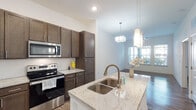 This single-floor residence presents a cohesive, modern design throughout its open layout. The kitchen serves as a central highlight, featuring rich dark-brown shaker cabinetry extending to the ceiling for generous storage, light granite countertops, and a full suite of stainless steel appliances including a range, over-the-range microwave, and refrigerator. A white-finished island with a matching granite surface and double undermount sink anchors the space, complemented by stylish pendant lighting overhead. The kitchen flows seamlessly into the living area, where hardwood-style flooring continues underfoot, a ceiling fan provides comfort, and a large window invites natural light and outdoor views.

The bathroom is well-appointed with a walk-in shower enclosed in glass, built-in wall shelving, a dark-wood vanity with a stone-topped single sink, and warm hardwood-style flooring. The tall ceilings lend an airy, open feel to the space. A separate bedroom offers hardwood-style flooring, a ceiling fan, and a window overlooking the outdoors. The 3D floor plan scan reveals an efficient, open-concept arrangement with clearly defined living, kitchen, and private areas connected by a practical, flowing layout.