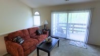 The living area of this single-floor space is designed to maximize natural light and openness. A large sliding glass door dressed with vertical blinds serves as the room's focal point, offering direct access to an outdoor balcony where lush greenery is visible, creating a pleasant indoor-outdoor connection. An arched accent window above a side console adds architectural character while allowing additional natural light to filter into the space. The neutral, light-toned walls complement the wall-to-wall carpeting, contributing to a warm and inviting atmosphere throughout the room. The layout comfortably accommodates a full-sized sofa and a coffee table, indicating a generous and functional living area well-suited for relaxing or entertaining. A side table with a lamp provides ambient lighting options for evening use. Overall, the space strikes a balance between comfort and practicality, with its thoughtful window placement ensuring the room feels bright and airy throughout the day while maintaining a cozy residential character.