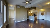 This single-floor space is designed with an open-concept layout that creates a fluid connection between the living area and kitchen. Rich, dark hardwood floors run throughout, adding warmth and a polished aesthetic to the interior. The living area is generously proportioned, featuring a ceiling fan with an integrated light fixture that provides both comfort and illumination. Earthy olive-toned accent walls add depth and character to the space, complementing the clean white trim and baseboards throughout.

The kitchen is thoughtfully designed with a raised bar-height peninsula that serves as a natural divider between the cooking and living areas. Pendant lights hang above the peninsula, casting focused task lighting over the dark countertops. Light wood cabinetry lines the kitchen walls, paired with stainless steel appliances including a built-in microwave. Under-cabinet lighting enhances both the functionality and ambiance of the cooking space. Recessed lighting and track lighting in the kitchen area ensure the space is well-lit throughout. The open sightlines from every angle make this layout ideal for entertaining and everyday living.