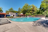 This expansive outdoor amenity area centers around a large, freeform swimming pool with clear turquoise water, accessible via stainless steel handrails at a gradual entry point. The pool is surrounded by a broad concrete pool deck offering ample space for lounging, lined with modern chaise lounge chairs and side tables arranged for comfort and convenience.

Framing the space are two distinctive red-stained wood pergolas that provide shaded gathering areas and add a warm architectural contrast to the neutral concrete hardscape. Adjacent to the pergolas is a cluster of accessory structures clad in blue and gray siding with a pitched shingle roof, which appear to house restroom or utility facilities, completing the self-contained nature of the amenity area.

The perimeter of the pool deck is enclosed by a low masonry wall with layered plantings of ornamental grasses, flowering shrubs, and mature deciduous and evergreen trees, creating a natural privacy screen and a lush, park-like atmosphere. The generous tree canopy and dense landscaping give the area a secluded, resort-like quality while allowing full exposure to natural sunlight throughout the day.

Overall, the space presents a well-maintained, thoughtfully designed outdoor retreat that balances open recreational space with shade, greenery, and architectural detail.