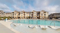 This community amenity space is designed for outdoor living and entertainment, centered around a generously sized swimming pool with a dedicated shallow lounge area equipped with built-in water features and step-in entry points. The pool deck is bordered by a wrought-iron fence and lined with lounge chairs, offering a resort-like atmosphere with views of the surrounding multi-story residential buildings.

Adjacent to the pool is a covered cabana structure that provides a shaded gathering area. The cabana features a warm wood-paneled ceiling with recessed lighting and a ceiling fan, complemented by light-toned shiplap-style walls and neutral tile flooring throughout. The space is furnished with ornate cast-iron bar-height tables and swivel chairs, as well as a round café-style table with matching seating, creating a versatile layout for both casual dining and social gatherings.

A built-in wet bar anchors one side of the cabana, featuring a dark granite countertop, a stainless steel sink, and a brick base surround that adds a classic, refined aesthetic. A window above the bar area allows natural light to filter in while maintaining views of the surrounding grounds. Stone-accented columns frame the open sides of the cabana, seamlessly connecting the covered area to the pool deck and broader outdoor environment.