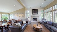 This single-floor community space is thoughtfully designed to foster comfort and social interaction. The main lounge area serves as the centerpiece, anchored by a striking floor-to-ceiling stone fireplace topped with a bold framed artwork piece and decorative topiaries along the mantel. Surrounding the fireplace, plush tufted leather sofas and patterned accent chairs are arranged atop an ornate area rug, creating an inviting conversational setting.

Large multi-pane windows line the walls, flooding the interior with natural light and offering pleasant views of mature trees and landscaped grounds. The soaring vaulted ceiling enhances the sense of openness throughout the lounge. A wall-mounted flat-screen television is positioned for easy viewing from the seating area.

A dedicated dining section features multiple round wooden tables with chairs, accented by a series of sculptural metallic wall art pieces illuminated by track lighting. This area benefits from its own generous window views overlooking a tranquil pond and greenery.

A corridor leads to additional functional areas, including a coffee station, further reinforcing the communal nature of the space. Consistent patterned carpeting and warm neutral tones throughout tie all areas together cohesively.