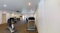 This fitness center offers a practical and well-organized space designed to support a full range of workout routines. The room features a carpeted floor that provides cushioning underfoot, making it comfortable for a variety of exercises. A textured white ceiling is fitted with recessed lighting and a ceiling fan, ensuring the space remains well-lit and properly ventilated during workouts. The walls are painted in a neutral off-white tone, contributing to a clean and open atmosphere. Equipment on offer includes a TRUE treadmill for cardio training, a multi-station strength training machine, a stair climber or elliptical, and additional fitness apparatus arranged throughout the room. Natural light filters in through windows along one wall, brightening the space during daytime hours. A wall clock keeps exercisers on schedule, while a posted notice board near the entrance provides relevant facility information. The overall layout is thoughtfully organized, allowing users to move freely between machines without feeling cramped. This is a functional and accessible fitness facility suitable for a wide range of fitness levels and training goals.