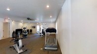 This fitness center offers a practical and well-organized space designed to support a full range of workout routines. The room features a carpeted floor that provides cushioning underfoot, making it comfortable for a variety of exercises. A textured white ceiling is fitted with recessed lighting and a ceiling fan, ensuring the space remains well-lit and properly ventilated during workouts. The walls are painted in a neutral off-white tone, contributing to a clean and open atmosphere. Equipment on offer includes a TRUE treadmill for cardio training, a multi-station strength training machine, a stair climber or elliptical, and additional fitness apparatus arranged throughout the room. Natural light filters in through windows along one wall, brightening the space during daytime hours. A wall clock keeps exercisers on schedule, while a posted notice board near the entrance provides relevant facility information. The overall layout is thoughtfully organized, allowing users to move freely between machines without feeling cramped. This is a functional and accessible fitness facility suitable for a wide range of fitness levels and training goals.