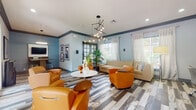 Located at 9051 SW 122nd Ave in Miami, FL, this well-appointed single-floor community space offers a thoughtfully designed interior that blends comfort and functionality. The main lounge area is anchored by a striking multi-bulb pendant chandelier that serves as a focal point, casting warm light over a mix of vibrant orange leather armchairs and a neutral sectional sofa arranged around a sleek circular white table. The walls are painted in a soothing blue-gray tone, complemented by crown molding and curated framed artwork that adds a refined, gallery-like quality to the space. Underfoot, a bold chevron-patterned mixed-tone wood-look flooring runs throughout, adding visual depth and texture. Large windows fitted with sheer drapes and horizontal blinds allow generous natural light to filter in, creating an airy ambiance. A dedicated work nook features a wall-mounted flat-screen TV, a wooden bar-height table with stools, and a circular pendant light — ideal for casual meetings or individual work sessions. Additional workstations with computer monitors are neatly arranged along one wall. Dual glass-panel entry doors open to the outdoor surroundings, further enhancing the connection to the exterior environment. Tropical greenery throughout adds a fresh, South Florida character to this vibrant and functional gathering space.