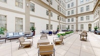 Located in the heart of Richmond, Virginia, this sophisticated open-air courtyard is enclosed on all sides by a stately multi-story neoclassical building featuring symmetrical rows of tall windows, crisp cream-colored facades, and refined architectural detailing. The courtyard is paved with large-format light stone tiles that lend a clean, expansive feel to the space, allowing natural light to pour in from above and illuminate the entire area throughout the day.

The space is thoughtfully furnished with an eclectic mix of seating arrangements, including woven rattan lounge chairs, cushioned sofas with blue upholstery, bistro-style tables with perforated metal chairs, and chaise lounges positioned for relaxation. These varied seating clusters are distributed across the courtyard, accommodating both intimate gatherings and larger social groups.

Raised planter beds filled with lush greenery, including hostas and other ornamental plants, add natural color and vibrancy against the neutral palette of the building. Overhead, strands of globe string lights are draped across the courtyard, creating a warm and inviting ambiance for evening use. At one end, a glass-enclosed interior space connects the courtyard to the building's interior. The overall atmosphere is polished, open, and ideal for both relaxed gatherings and professional events.