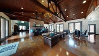 This single-floor clubhouse is a well-appointed communal facility that blends rustic charm with modern functionality. The centerpiece of the space is a grand open lounge area distinguished by dramatically vaulted, wood-paneled ceilings supported by exposed heavy timber beams. A striking wrought-iron chandelier hangs overhead, casting warm light across the polished stained concrete floors that run throughout. A full-height brick fireplace anchors one wall, framed by a wooden mantel and accented with decorative turquoise pieces, creating an inviting focal point. Comfortable leather Chesterfield sofas and armchairs are arranged throughout the lounge, complemented by tufted ottomans and teal accent pillows. Large French doors and plantation-shuttered windows allow generous natural light to flow in while offering views of the surrounding exterior. A dedicated dining area features a large wooden table with ample seating, pendant lighting, and direct outdoor access via French doors. The full galley-style kitchen is equipped with dark cabinetry, granite countertops, stainless steel and black appliances, and a secondary prep area with additional counter space. A business center provides multiple computer workstations for residents' use. The overall ambiance is warm, welcoming, and richly detailed, combining rustic architectural elements with comfortable, functional amenities.