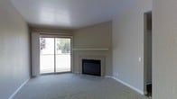 This single-floor space centers around a comfortable living area defined by warm, neutral-toned walls and plush carpeted flooring. A standout feature is the tile-surround fireplace, positioned against the main wall and topped with a clean white mantel shelf, providing both a functional heating element and a natural focal point for the room. Adjacent to the fireplace, a sliding glass door floods the space with natural light and offers views of outdoor greenery, creating a bright and airy atmosphere. The door leads to what appears to be a balcony or patio area, bordered by a white railing. The textured ceiling adds subtle depth to the room, while white baseboards provide crisp contrast against the carpeting and walls. The overall design is understated and clean, with a simple, functional layout that offers a versatile canvas for furnishing and personalization. The combination of natural light, a fireplace, and outdoor access contributes to an inviting and livable ambiance throughout the space.
