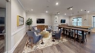 This well-appointed single-floor amenity space is designed to offer a versatile and inviting environment for gathering, relaxing, and entertaining. The layout flows seamlessly between distinct functional zones, each thoughtfully designed with modern finishes and cohesive décor throughout.

The full kitchen is outfitted with dark espresso cabinetry, white quartz countertops, and a full suite of stainless steel appliances, including a refrigerator, range, dishwasher, and microwave. A decorative mosaic tile backsplash adds visual interest, while light-toned tile flooring enhances the clean, contemporary aesthetic. The kitchen offers ample counter and storage space, making it well-suited for catering or communal use.

The lounge area is a warm and welcoming space featuring rich hardwood-style flooring, crown molding, and textured accent wallpaper. Comfortable blue upholstered chairs are arranged around a woven rattan coffee table atop a large patterned area rug, creating an ideal setting for casual conversation. A wall-mounted flat-screen television serves as a focal point, complemented by framed artwork and tasteful décor.

Adjacent to the lounge, a billiards table anchors a game area, while a bar-height communal table with industrial-style stools provides additional gathering space. Recessed and track lighting throughout ensure the space is well-lit and inviting at any time of day.