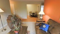 This single-floor residence offers a well-connected layout that seamlessly integrates the living and dining areas. The living room serves as the central gathering space, featuring warm wood-look flooring partially covered by a patterned area rug, which adds texture and visual interest underfoot. A bold terracotta-orange accent wall provides a striking focal point, complementing the neutral taupe tones found throughout the rest of the space. The room accommodates comfortable seating, including a plush brown sofa adorned with decorative pillows, alongside a dark wood coffee table and matching end table with a classic table lamp. A large potted plant near the seating area introduces a natural, lively element to the interior. An open passageway leads directly into the adjacent dining area, which features a dark wood circular dining table surrounded by upholstered chairs — ideal for shared meals. Natural light filters in through a window with vertical blinds visible in the background, brightening the space. The overall ambiance is warm and inviting, with a casual, livable aesthetic that balances comfort and functionality throughout.