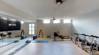 This fitness studio is designed to support a wide range of workout routines in a clean, modern setting. The space features light wood-style flooring throughout, which provides a neutral and easy-to-maintain surface suitable for both equipment use and floor exercises. A row of high horizontal windows lines the upper portion of the walls, allowing ample natural light to fill the room while maintaining privacy. Ceiling fans help maintain comfortable airflow during workouts.

The studio is equipped with a variety of fitness equipment, including a rowing machine, a stationary bike, and a step platform, catering to both cardio and strength training needs. A dedicated dumbbell rack holds a full range of colorful free weights, making strength-based exercises convenient and accessible. Yoga mats and balance tools such as a BOSU ball and foam step blocks are available for stretching, balance training, and low-impact workouts.

A large floor-to-ceiling mirror is mounted along one wall, enhancing the sense of space and allowing users to monitor their form during exercise. The walls are finished in a soft, neutral gray tone that creates a calm and focused atmosphere. The overall layout is open and uncluttered, offering generous room to move freely between equipment and designated exercise zones.