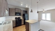 This single-story space showcases an open-concept kitchen that serves as a central hub of the layout. The kitchen is outfitted with dark taupe cabinetry offering generous storage, complemented by a classic white subway tile backsplash that adds a clean, timeless contrast. Black appliances, including a range, microwave, and refrigerator, provide a sleek, cohesive look throughout the cooking area. A built-in wine rack adds a thoughtful design detail above the upper cabinetry. The granite-topped kitchen island acts as a natural dividing point between the kitchen and the adjoining living area, and is illuminated by two industrial-style pendant lights with metallic finishes, adding character and focused task lighting. Track lighting along the kitchen ceiling further enhances functionality. The flooring transitions from warm hardwood-style planks in the kitchen to plush carpeting in the living area, clearly delineating the two spaces. A doorway visible in the background hints at additional natural light entering the space, contributing to an open and airy atmosphere throughout this well-appointed single-floor layout.