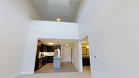 This 1-bedroom loft is a two-story residence that makes a striking impression with its soaring double-height ceilings, which flood the main living area with a sense of openness and volume. The open-concept layout seamlessly connects the living space to a well-appointed kitchen, which features dark cabinetry, stainless steel appliances, and a central island with light stone countertops — perfect for both meal prep and casual dining. A pendant light above the island adds a refined touch to the space. The clean white walls throughout the main level enhance the bright, airy ambiance, while light-toned flooring provides a warm, neutral foundation. A glimpse into the bathroom reveals coordinating dark cabinetry with ample counter space, maintaining a consistent modern aesthetic throughout the unit. The entryway, framed by a classic white door, offers a welcoming transition into the home. The two-floor configuration provides distinct zones for living and sleeping, offering both functionality and privacy. Overall, this loft combines contemporary design with practical layout choices, making it an efficient and visually compelling living space.