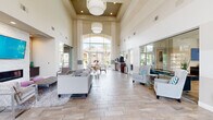 This single-floor community clubhouse is designed to impress from the moment you step inside. The grand main hall commands attention with its dramatic double-height ceilings, oversized pendant light fixtures, and floor-to-ceiling arched windows that flood the space with natural light and offer sweeping views of palm-lined outdoor surroundings. Wood-look tile flooring flows seamlessly throughout, tying the various areas together in a cohesive, contemporary aesthetic.

The main lounge area provides generous seating with plush sofas and accent chairs arranged around a sleek linear fireplace framed in white stacked stone, with a mounted flat-screen TV and decorative purple accent niches flanking each side. A separate, more intimate lounge features a geometric accent wall, sectional seating, and an additional mounted TV. 

A dedicated leasing office with glass partition walls offers both transparency and privacy, while a well-appointed fitness center is equipped with cardio machines, strength equipment, floor-to-ceiling mirrors, and mounted TVs. The community kitchen stands out with a professional-grade stainless steel range, mosaic tile backsplash, and stone countertops. Glass entry doors with adjacent potted greenery complete the welcoming, resort-style atmosphere of this thoughtfully designed space.