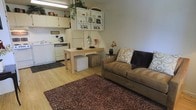 This single-floor space offers an efficient and well-organized open-concept layout that integrates a living area and kitchen into one cohesive environment. The light wood laminate flooring runs throughout, lending warmth and visual continuity to the space. A plush, dark shag area rug anchors the living zone, where a comfortable tan sofa is accented with decorative throw pillows, creating an inviting place to relax. A simple, light-toned dining table paired with cube ottomans serves as a versatile surface for both meals and casual use. The kitchen is positioned along one wall and features white cabinetry, a full-size refrigerator, dishwasher, and a range, providing essential appliances in a compact footprint. A dark floor mat defines the kitchen floor area. Above the kitchen, a wall-mounted open shelving unit displays decorative items and small plants, adding a personal touch and making excellent use of vertical space. Trailing greenery and small potted plants throughout contribute a natural, lived-in ambiance. The neutral color palette of whites, beiges, and browns creates a calm, cohesive atmosphere throughout the space.
