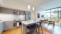 Located in Tampa, FL, this thoughtfully designed residence offers a seamless blend of contemporary architecture and refined interior finishes. The open-concept kitchen is a standout, featuring sleek grey and blue cabinetry, white subway tile backsplash, stainless steel appliances, and a central island with bar seating, all illuminated by elegant pendant lighting that hangs from soaring ceilings. Warm hardwood flooring flows throughout the living areas, connecting the kitchen to a welcoming dining space and a bright living room anchored by dramatic floor-to-ceiling windows. These expansive glass panels flood the space with natural light and frame sweeping views of the surrounding Tampa cityscape and greenery. The bedrooms continue the home's calm, neutral aesthetic, with large windows offering city views and generous natural light. The bathrooms are finished with light wood vanities, white quartz countertops, large-format tile flooring, and both a glass-enclosed walk-in shower and a separate soaking tub option. Throughout the residence, high ceilings and an airy, open layout create a sense of spaciousness, while the warm tones and curated design elements contribute to an inviting and modern urban ambiance.