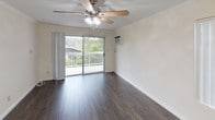 This single-floor unit offers a practical and comfortable layout with distinct living, sleeping, and bathroom areas. The open living and dining area features dark hardwood-style flooring, crown molding, and a ceiling fan with integrated lighting. A large window with vertical blinds allows natural light to fill the space, while a sliding glass door provides direct access to a balcony overlooking lush, tree-lined surroundings — a notable feature that enhances the overall ambiance. The kitchen sits adjacent to the living area and is equipped with white cabinetry, dark countertops, and white appliances including a refrigerator and range.

The bedrooms are carpeted in a neutral tone and share a consistent design language with crown molding and ceiling fans with lights. Each room includes windows offering outdoor views, and at least one bedroom features a wall-mounted air conditioning unit for individualized climate control. The bathroom is finished with warm-toned tile flooring and includes a full-size bathtub and shower combination enclosed by a sleek sliding glass panel. The tub surround features large-format tile with a decorative mosaic accent border, complemented by dark granite-topped cabinetry. A small frosted window provides ventilation and privacy. Throughout the unit, the design emphasizes clean, neutral tones and functional finishes.