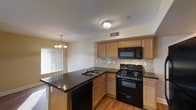 This two-story residence offers a thoughtfully designed interior highlighted by an open-concept kitchen that serves as a central hub of the living space. The kitchen is outfitted with medium-toned wood cabinetry providing ample storage, complemented by dark granite-style countertops that add a striking contrast. A peninsula with an integrated sink and built-in dishwasher extends into the space, creating a natural divide between the kitchen and adjacent dining or living area. Black appliances, including a gas range, built-in microwave, and dishwasher, provide a cohesive, modern aesthetic throughout. A mosaic tile backsplash adds texture and visual interest along the cooking wall. Warm hardwood-style flooring runs throughout the main level, contributing to an inviting atmosphere. Natural light filters in through a window fitted with horizontal blinds, brightening the space. Overhead, a flush-mount ceiling fixture provides general illumination, while a classic chandelier above the dining area adds a decorative touch. The two-floor layout offers separation between living and private spaces, providing functional versatility for everyday living.