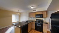 This two-story residence offers a thoughtfully designed interior highlighted by an open-concept kitchen that serves as a central hub of the living space. The kitchen is outfitted with medium-toned wood cabinetry providing ample storage, complemented by dark granite-style countertops that add a striking contrast. A peninsula with an integrated sink and built-in dishwasher extends into the space, creating a natural divide between the kitchen and adjacent dining or living area. Black appliances, including a gas range, built-in microwave, and dishwasher, provide a cohesive, modern aesthetic throughout. A mosaic tile backsplash adds texture and visual interest along the cooking wall. Warm hardwood-style flooring runs throughout the main level, contributing to an inviting atmosphere. Natural light filters in through a window fitted with horizontal blinds, brightening the space. Overhead, a flush-mount ceiling fixture provides general illumination, while a classic chandelier above the dining area adds a decorative touch. The two-floor layout offers separation between living and private spaces, providing functional versatility for everyday living.