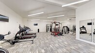 This fitness center in Russellville, Arkansas, offers a generously sized, single-floor layout designed to accommodate a comprehensive range of workout activities. The space features soaring vaulted ceilings that enhance the open, airy atmosphere, while rows of suspended linear LED fixtures provide bright, uniform illumination throughout. The flooring consists of durable, patterned carpet tiles in neutral grey tones, contributing to both comfort underfoot and a clean, modern aesthetic.

The cardio zone includes multiple treadmills and an Endurance-brand rowing machine, each positioned along one side of the room for an organized, efficient use of space. Wall-mounted flat-screen televisions are strategically placed throughout, offering entertainment during workouts. The strength training area features a robust power rack with an attached bench, a multi-function cable machine, a tiered dumbbell rack stocked with a full range of weights, and an adjustable workout bench. Full-length mirrors line portions of the walls, aiding in form correction and visually expanding the space.

The layout allows ample open floor space between equipment zones, supporting safe movement and potential stretching or floor exercises. A glass-panel entry door offers visibility into the space. The overall design is clean, functional, and well-organized, reflecting a professional fitness environment.