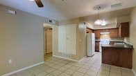 This single-story space offers a straightforward, functional layout with an open flow between the main living area and kitchen. Light beige tile flooring runs consistently throughout, lending a cohesive and easy-to-maintain foundation across the space. The neutral-toned walls create a clean, versatile backdrop suitable for a variety of interior styles.

The kitchen is positioned toward one end of the space and features warm wood-toned cabinetry providing generous upper and lower storage. A white refrigerator and white range are included appliances, complemented by a dark countertop that adds a subtle contrast. A pendant light fixture hangs overhead, offering focused task lighting above the kitchen area.

Adjacent to the kitchen, the main living area is illuminated by a ceiling fan, supporting both air circulation and overhead lighting. A set of double closet doors provides additional built-in storage, a practical feature within the main living zone. A hallway leads toward additional rooms, including what appears to be a bedroom or secondary space with its own door. HVAC vents are visible throughout, indicating a central air system. Overall, the space presents a clean, practical interior with functional amenities.
