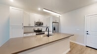 This single-floor residence at 4665 Whitfield Ave in Sarasota, Florida offers a thoughtfully designed interior with modern finishes throughout. The kitchen serves as a standout feature, showcasing crisp white shaker cabinetry, dark countertops, and a striking elongated pendant light above a generous island with an integrated sink and matte black faucet. A geometric white tile backsplash adds visual interest, while stainless steel appliances — including a refrigerator with ice and water dispenser, electric range, and over-the-range microwave — complete the space. Light wood-look flooring runs through the main living areas, contributing to a bright, airy atmosphere. The living area benefits from natural light through multiple windows and a sliding glass door, complemented by a ceiling fan overhead. Bedrooms are finished with plush neutral carpet, ceiling fans, and window blinds that allow for natural light while maintaining privacy. The bathroom features a clean, white-tiled tub and shower surround with a timeless design. A dedicated laundry room accommodates full-size washer and dryer units with overhead shelving for added storage. The overall design palette of soft neutrals, white walls, and modern hardware creates a cohesive, contemporary ambiance throughout.